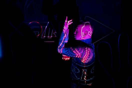 A man with neon-lit tattoos poses in a cyberpunk environment, surrounded by colorful lights.